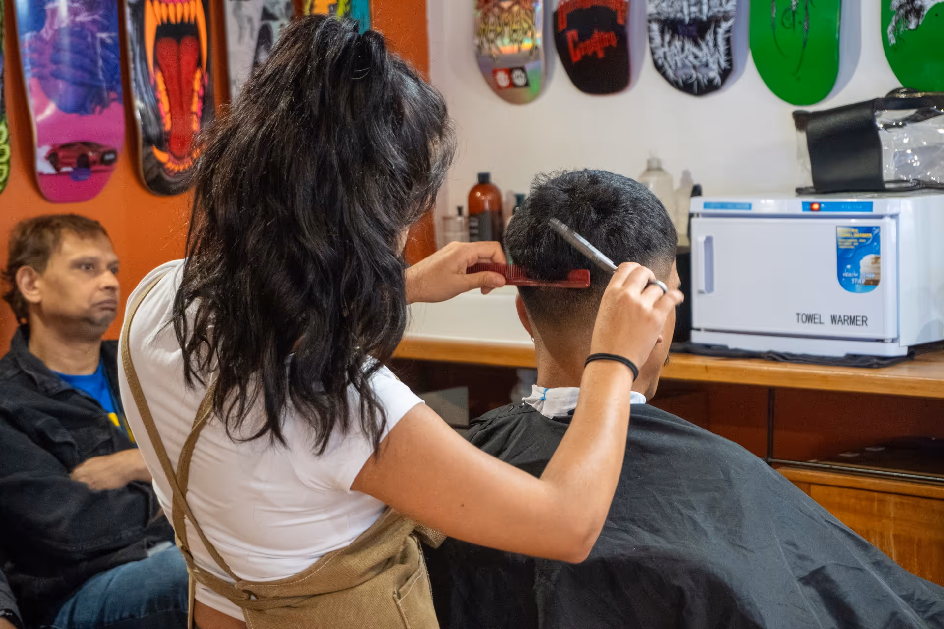 Barber with long dark hair cutting a male customer's short hair in a shop decorated with colorful skateboard decks.